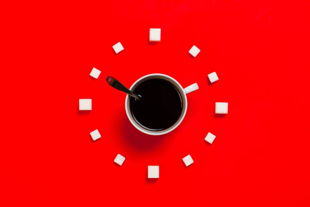 coffee cup on red background in the middle of sugar cubes marking points of a clock. Spoon in cup points to 10 oc lcock, cup handle to 2 oc clock