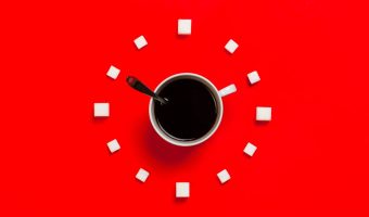 coffee cup on red background in the middle of sugar cubes marking points of a clock. Spoon in cup points to 10 oc lcock, cup handle to 2 oc clock