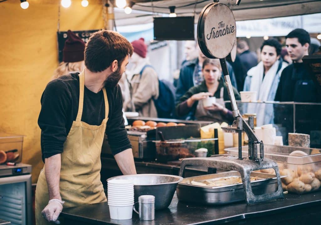 Man Standing in Front of Bowl in busy cafe and Looking Towards Left at his customers