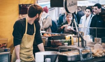 Man Standing in Front of Bowl in busy cafe and Looking Towards Left at his customers